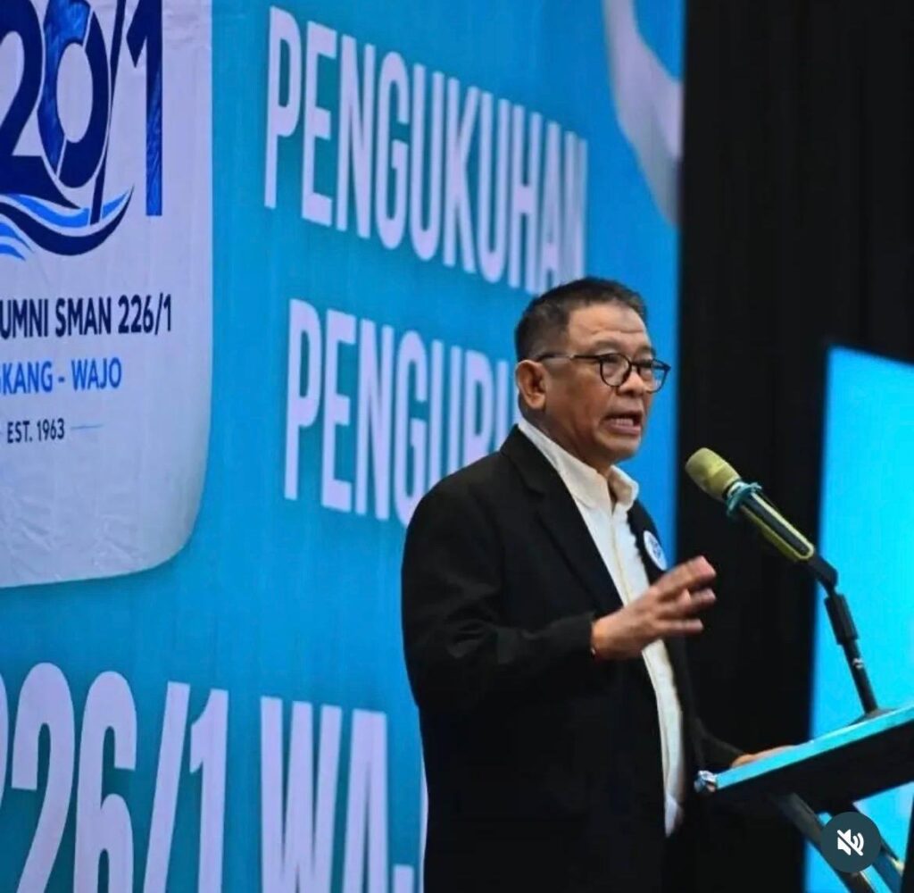 Man in a suit speaking at a podium on a stage with a large blue banner in the background reading pengukuhan pengurus